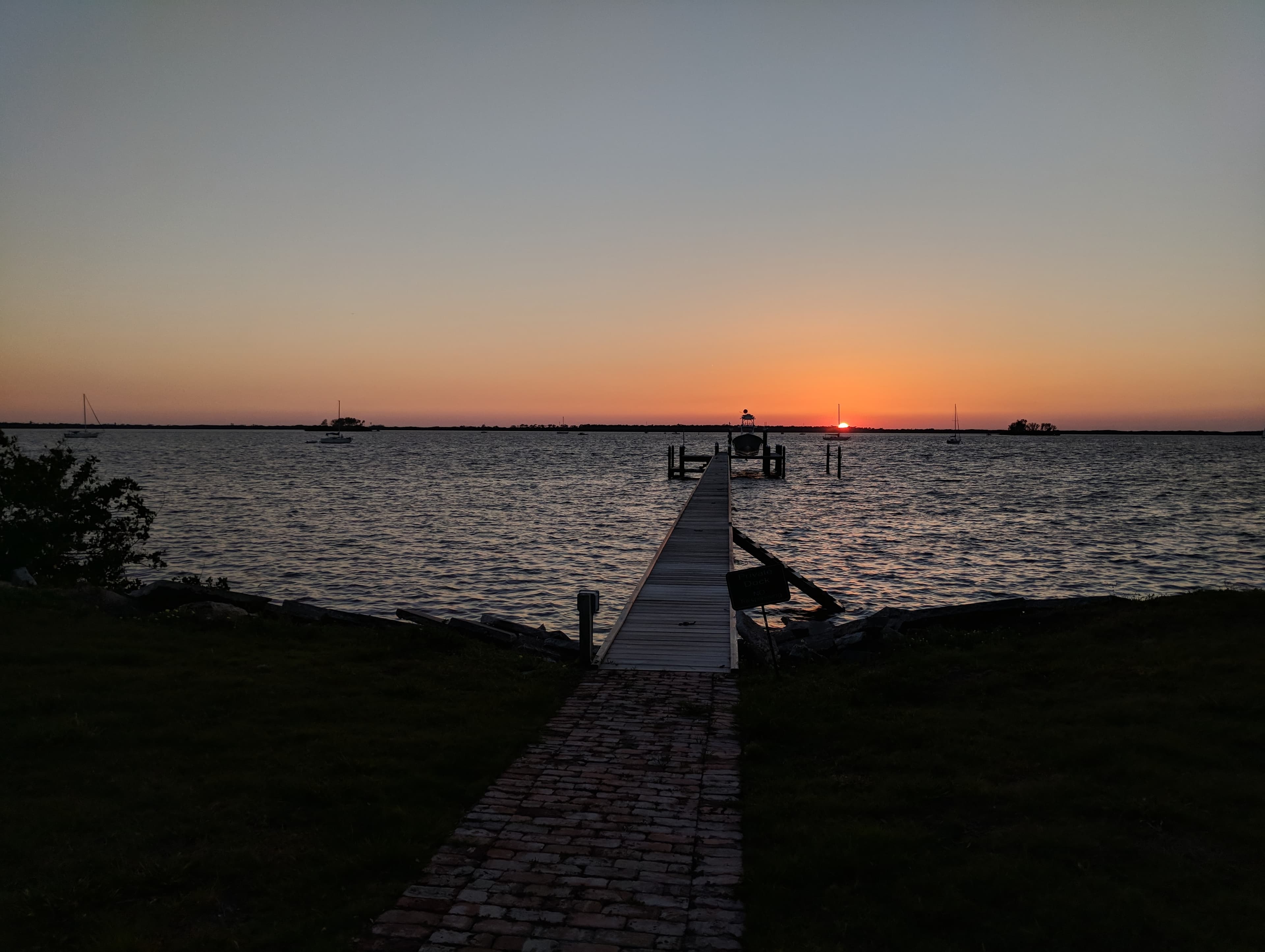 A wooden dock stretching into the bay at sunset, with sailboats on the water
