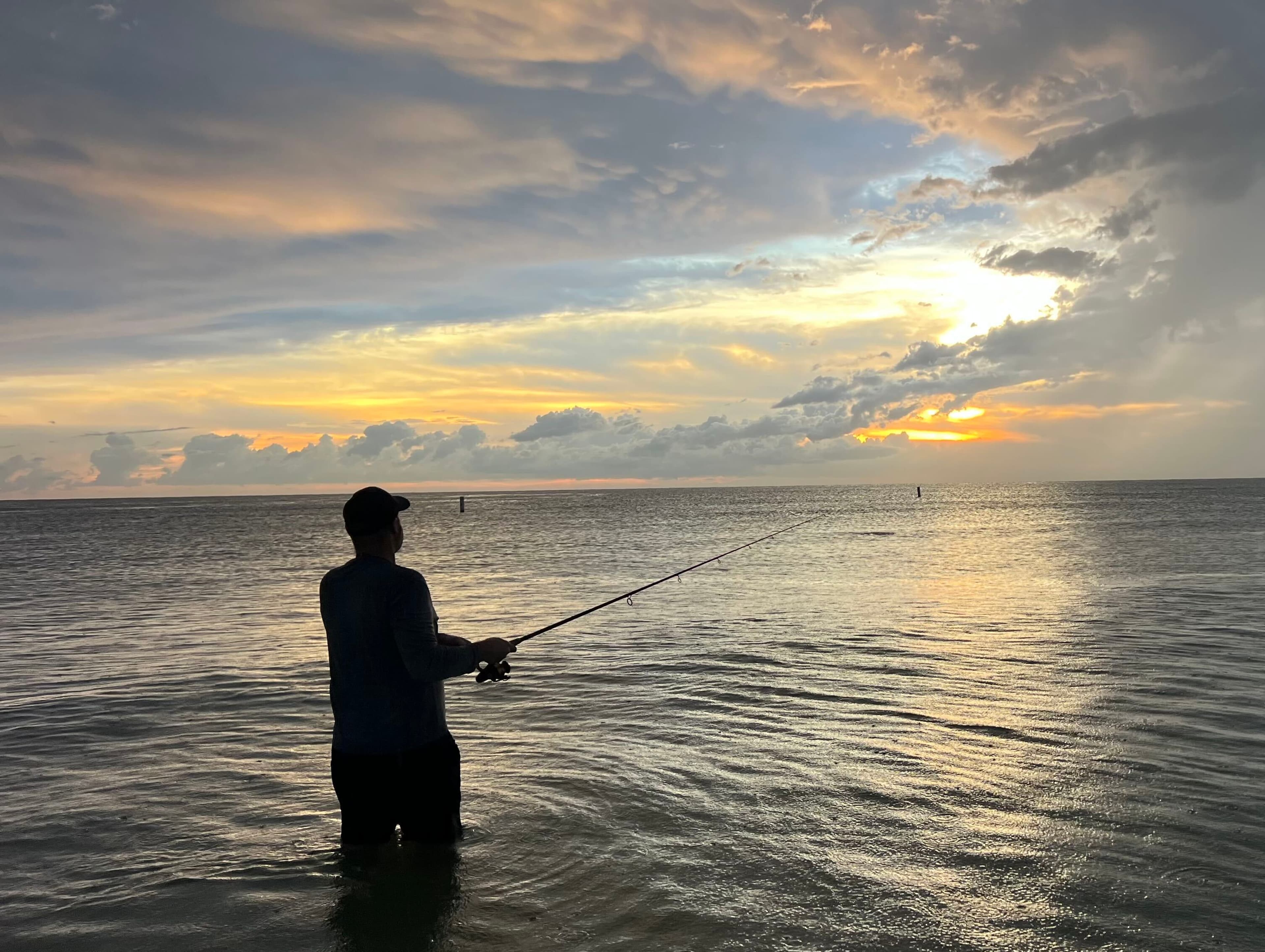 An angler wading in shallow water and fishing beneath a cloudy sunset
